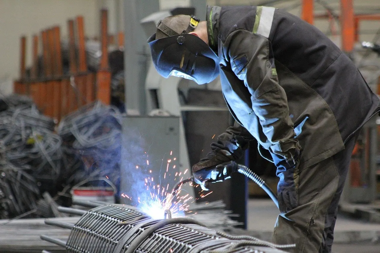 Schweißer arbeitet mit Schutzschild und funkensprühender Flamme an einem Metallgitter in einer Werkhalle.