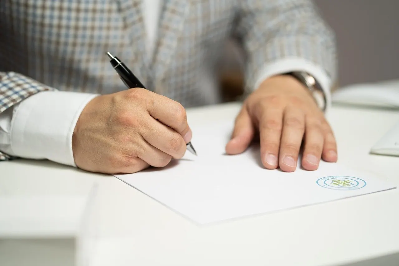 Person im karierten Sakko unterschreibt ein Dokument mit Stift an einem weißen Tisch, mit Siegel-Stempel