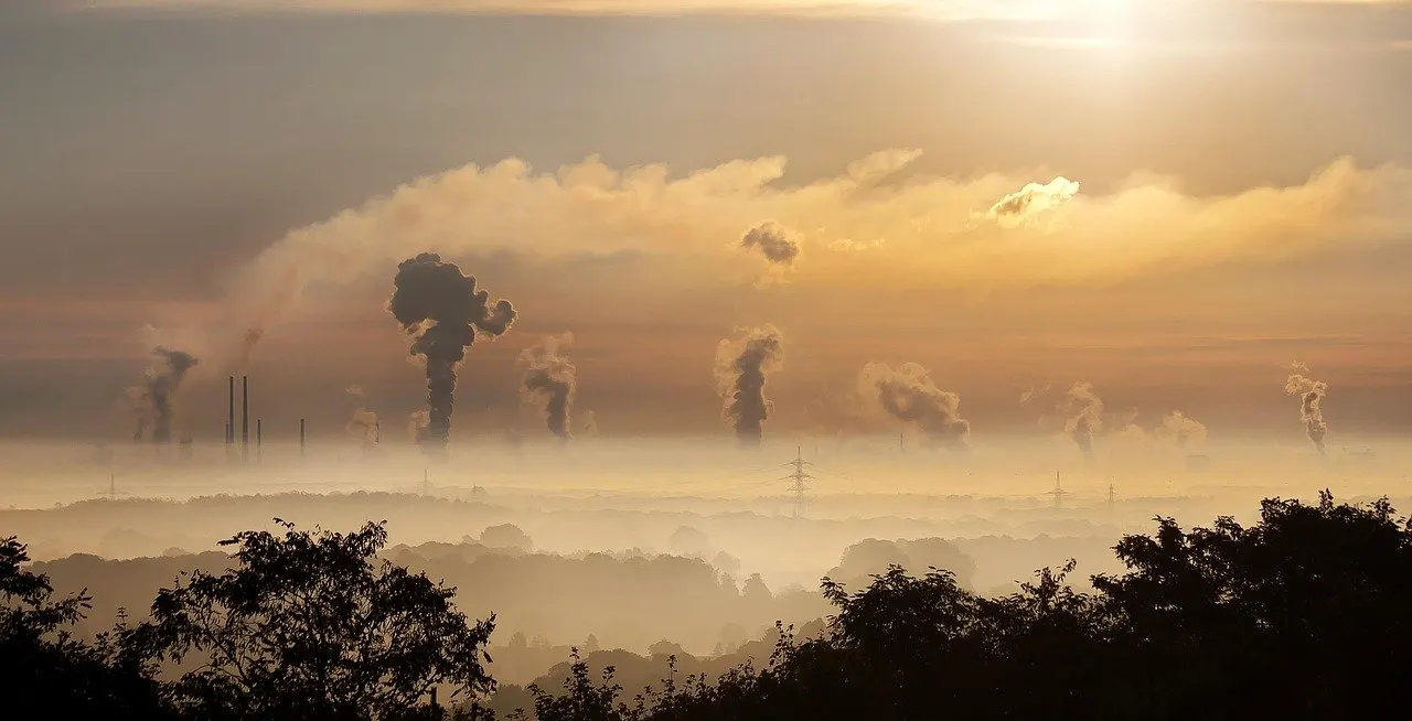 Morgendunst über einer Landschaft mit mehreren aufsteigenden Rauchfahnen von Schornsteinen, Sonnenlicht im Hintergrund.